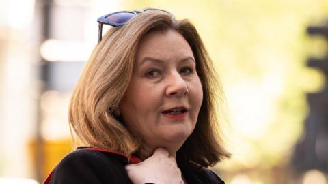 Teacher Rebecca Richardson has shoulder-length blonde/brown hair. She is wearing a black top and has her hand on a red bag strap and has sunglasses on her head. The background behind her is blurred.
