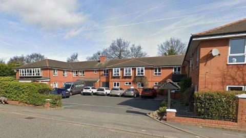 The exterior of the Aaron Court Care Home on a clear day with hedges at the side of the entrance to the car park and building.