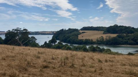 Dry, sun-scorched grassland slopes down towards a broad river estuary bordered by trees and rolling fields. A few small boats are visible on the water in the distance. Above, a vivid blue sky is filled with dramatic streaks and swathes of white cloud, contrasting with the dry, golden-brown landscape below.
