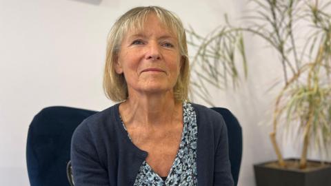 Dr Sian Wareing-Jones sat on a blue arm chair. She is wearing a blue cardigan and blue floral dress. The walls around her are white and there is a plant behind her. 