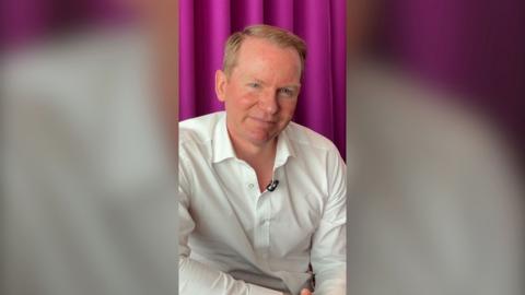 A man, Charlie Nunn, wears a white shirt. He has short red hair and sits in front of a purple curtain.