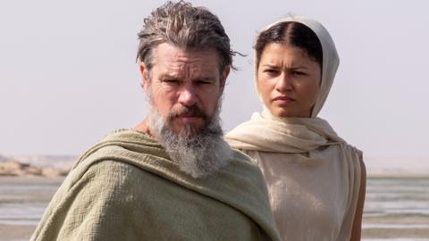 Matt Damon, with a long grey beard and wearing a light brown shroud, and Zendaya, whose brown hair is covered by a beige scarf to match her beige dress, portray the hero Odysseus and the goddess Athena in the film The Odyssey