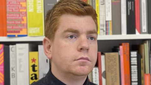 A man with ginger hair staring forward in front of a book case.