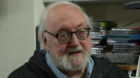 Martin Shipton, journalist, seated and wearing a checked shirt and a gilet, at his home in Cardiff