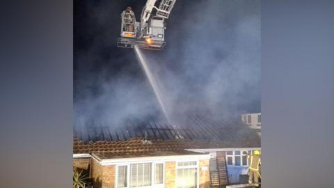 A firefighter on an aerial ladder platform using a water hose on a bungalow which has a burnt roof. 