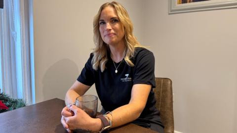 Martell Blackford sat at a table with a coffee in a glass mug. She has blonde air and is smiling. She is wearing a necklace and a black t-shirt that says 'Jacob's Table'. She is wearing a watch and some bracelets. She has a tattoo on her left hand. 