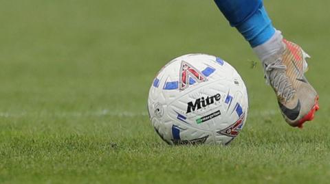 A National League football with 'HUFC' written on it being kicked by a white boot wearing a blue sock