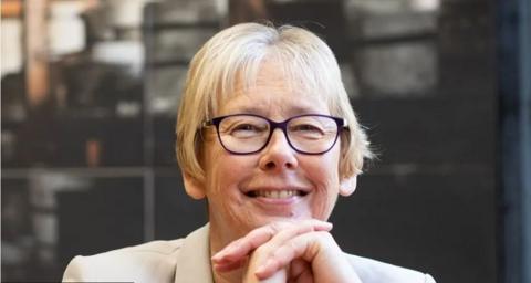 A woman with short blonde hair smiles at the camera. She wears dark blue rimmed glasses and has her chin balanced on her hands which have her fingers interlocked.