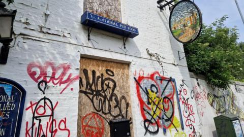 A close-up of the boarded up front door of a white building that used to be a pub. It has lots of colourful graffiti tags all over it.