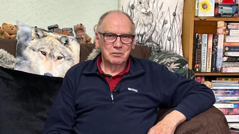 Brain Aves looking to the camera with a straight face. He is sitting on a brown sofa and is wearing blue jeans, a blue pullover and red shirt. There are various cushions on the sofa, including one that features a photo of a wolf, and cuddly toys above it.. There is a thin bookshelf to the right of him which is stacked with books.