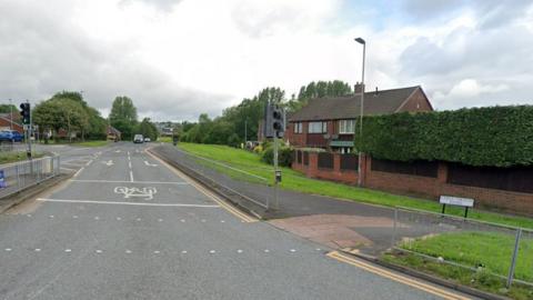 A road junction, with a sign reading Chell Heath Road on a grass verge. There is a light-controlled crossing at the junction and cars in the distance as well as houses either side.