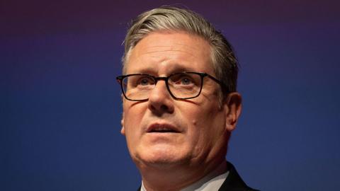 Sir Keir stood at a lectern at Welsh Labour conference. He is wearing a black suit, white shirt and black glasses.