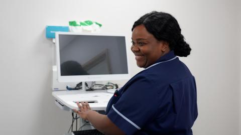 A nurse in a navy uniform operates a computer workstation inside a clinical room, with medical equipment and cables visible on the unit. The nurse is smiling.