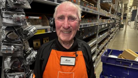 An older male employee stands in the electrical aisle of a B&Q store. He is smiling, and has grey hair. He's wearing a black shirt with an orange apron over the top, and his name badge says 'Ron'. There are blue crates behind him, and electrical equipment hung up to his left.