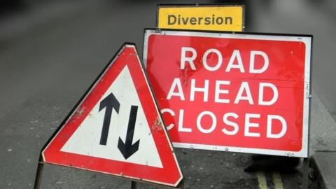 A red road ahead closed sign with another yellow diversion sign behind it