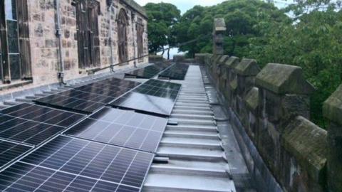 A long row of solar panels on the roof of a church behind a stone parapet  