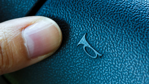 A close-up of someone's thumb on the dashboard button used to sound the horn. The black plastic button, which has a faux-leather pattern, has the symbol of a trumpet-like horn on it.