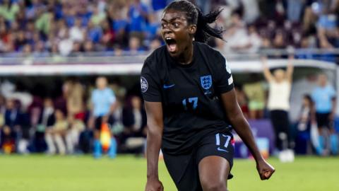 Michelle Agyemang in a dark England top, shouting and running on a football pitch.