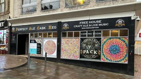 The entrance to a bar in Wolverhampton city centre. The front of the building in painted black and has a white and gold sign that reads "Perfect Ale Cask Keg". The windows have kaleidoscopic prints covering them as well as the bar's logo which is a grey and gold wolf.