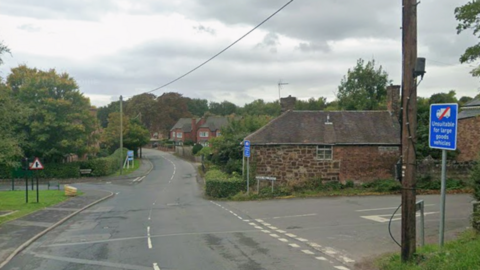 A road junction with a wooden pole in the foreground and a number of brown brick houses and trees on the far side