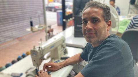 Sabur Kirimi wearing a blue tshirt and looking at the camera is sat in front of a sewing machine