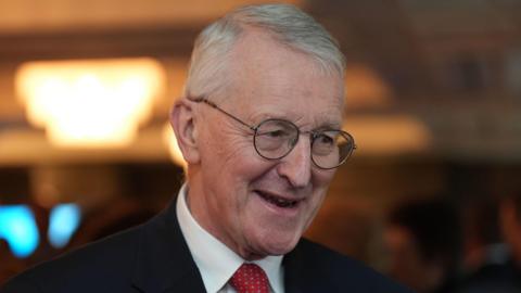 Hilary Benn smiling. He wears a black suit, white shirt and red tie. He has grey hair.