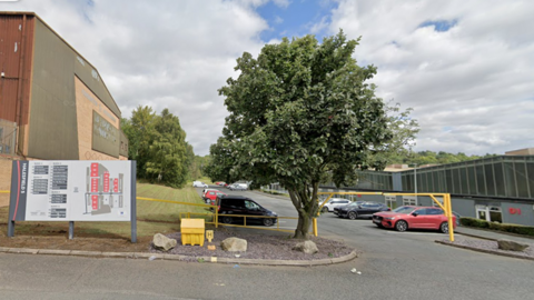 The entrance to a factory car park on an industrial estate. There is a big tree next to a yellow arch that cars drive under. There are factories on the left and right of the car park