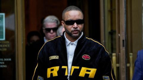 Stylist Deonte Nash, wearing a black and yellow jacket, blue shirt underneath and sunglasses, exits the courthouse. 