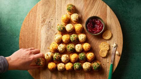 A circular chopping board sitting on a green table, on the chopping board there are rows of cheeseballs, lined up to resemble a Christmas tree. 