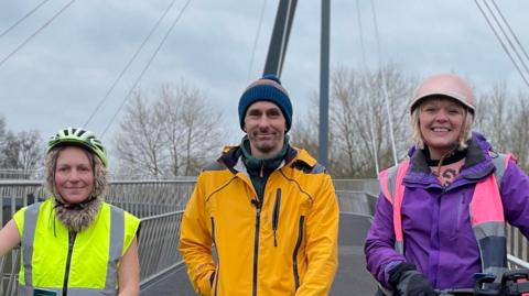 Three people - a woman wearing a hi-vis and black trousers and yellow cycle helmet, a man wearing an orange jacket, dark blue jeans and a beanie, and a woman wearing a purple coat, pink and grey hi-vis, light blue jeans and pink cycle helmet - stand next to their bikes on the Kepax footbridge, with temporary barriers either side of them.