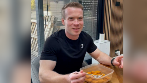 Man eating fish fingers out of a tupperware. He sits at a wooden table and wears a black t-shirt.