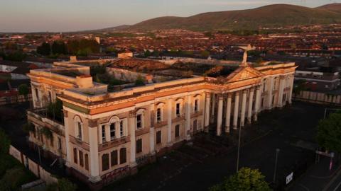 A still image taken from aerial drone footage of the former Crumlin Road Courthouse in north Belfast.  The large Victorial building has a Palladian portico but most of its roof is missing and there are scorch marks from previous arson attacks. 
