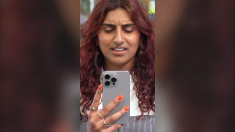 Woman with red hair holds a silver phone up to her face and looks confused