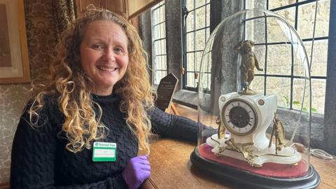 Faye Rason is smiling at the camera and her left arm is resting on a wooden  window shelf which is holding a clock in a glass dome about 0.5m (1ft) high. The clock is carved from a pale marble-type substance and has a white and gold-coloured face in which the mechanism can be seen. There is a gold-coloured sculpture of a cherub on the top of it and some symbolic sculptures below, one of which looks like an easel. 