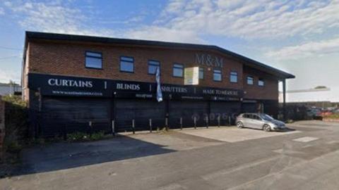 A two‑storey commercial building made of red brick, positioned along a wide paved road. The building has a slightly sloped roof and several rectangular windows evenly spaced along the upper floor. Across the ground floor, large black shuttered fronts display prominent white lettering that reads “CURTAINS”, “BLINDS”, “SHUTTERS”, and “MADE TO MEASURE”.