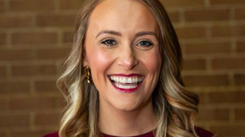 A woman with long blonde hair and gold earrings smiles at the camera