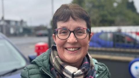 Karen McKenna - a woman with short brown hair smiles at the camera. She has a  pair of colourful square framed glasses on, as well as a plaid scarf and a green puffer coat on. She is standing in front of two parked blue cars and the background is blurred.