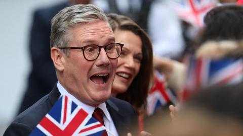 British Prime Minister Keir Starmer and his wife Victoria Starmer greet supporters outside downing street. Both people are smiling amid waving union flags.