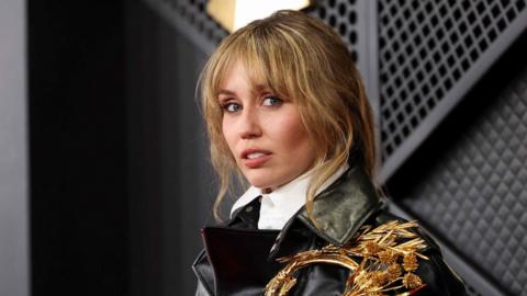 Miley Cyrus looks to the side of the camera wearing a black leather jacket and white shirt. She has brown/blonde hair with a pony-tail hairstyle.