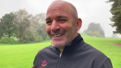 A man smiles while standing on a golf course in a black golfing jacket