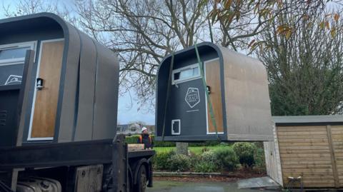 Two grey pods being delivered - one is being craned from a lorry to the ground. Each has a small window and brown door with keypad and Amazing Grace Spaces logo on the front.