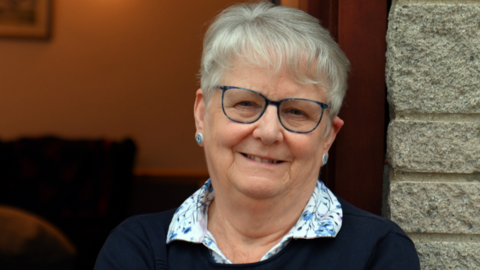 A lady in her 70s smiles at the camera, wearing blue-framed glasses and two blue button earrings. She wears a white blouse with blue cornflowers on the collar and a navy jumper over the top.