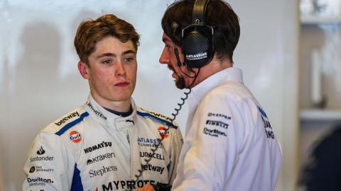 Luke Browning in the garage during F1 testing with Williams Racing, wearing a largely white race suit featuring several sponsor logos