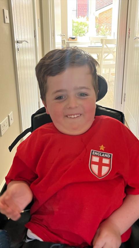 Young boy sat in wheelchair smiling wearing a red England foootball shirt