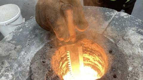 A person with protective gloves on holds a knife inside a circular furnace which is below a concrete chamber. The knife has a long serrated blade and is being held just above the blazing heat of the fire.