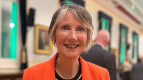 Fiona is wearing a bright orange blazer and a black top stands indoors at what appears to be a formal or social event. She has a name badge pinned to the blazer and a pearl-style necklace. The background shows a large hall with wooden flooring, blue chairs arranged in rows, tall windows with dark curtains, framed pictures on the walls, and several other attendees minglin