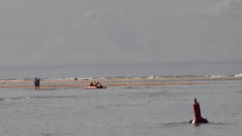 Two people on a sandbank in the middle distance as a lifeboat approaches. The is water between two separate areas of sandbank. A red buoy is floating in the foreground on the right.