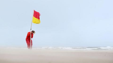 Lifeguard securing a red and yellow flag on a deserted beach with sand whipped up by strong winds 