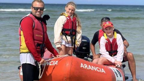 Two girls are wearing life jackets and are aboard a blow-up boat. The jackets are red. Annie is wearing a red hat. There are two adults next to them keeping the boat steady.