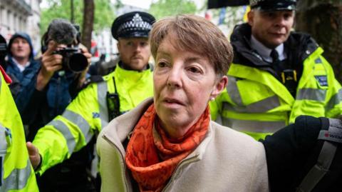 Former Post Office CEO Paula Vennells, wearing an orange scarf and beige coat and flanked by police officers in yellow high-vis coats, arrives to give evidence at the Horizon IT public inquiry on Wednesday, 22 May 2024. 